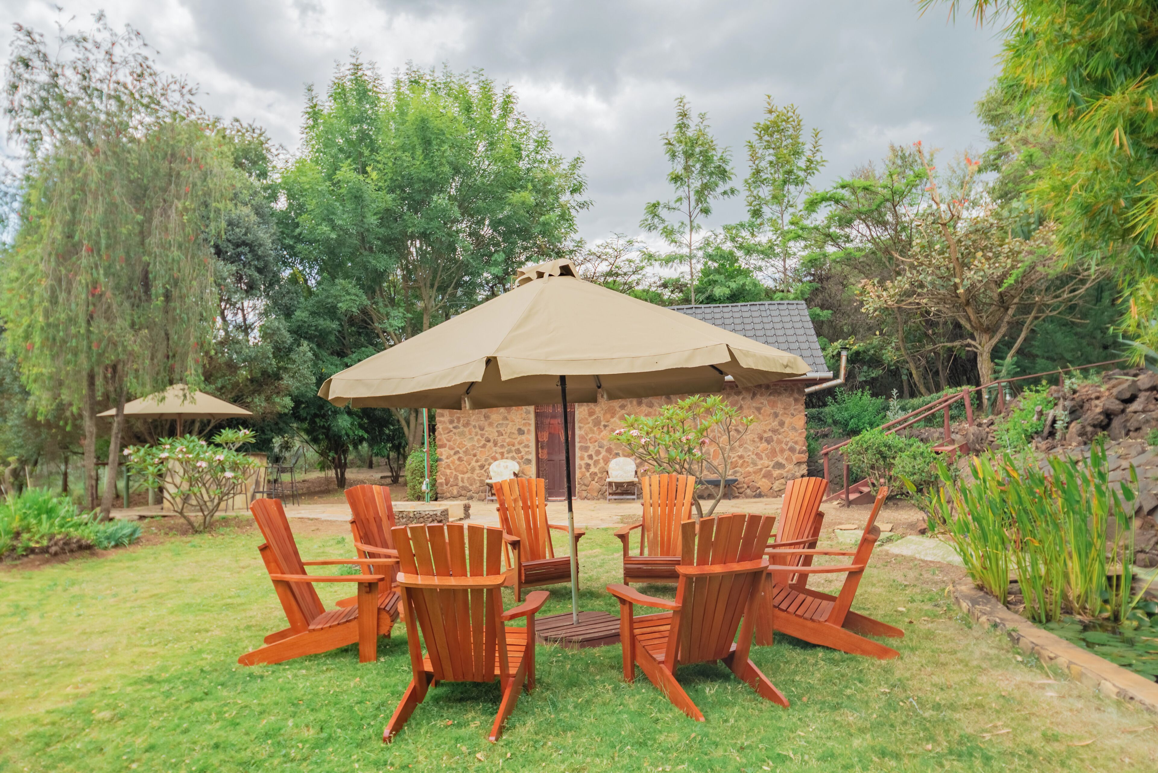 A patio area with wooden chairs and a large umbrella, surrounded by greenery and a stone building.