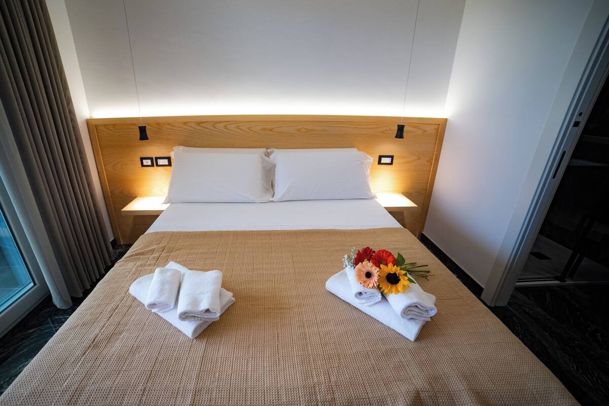 A hotel room with a bed, two bedside lamps, a window with blinds, and a wooden headboard.