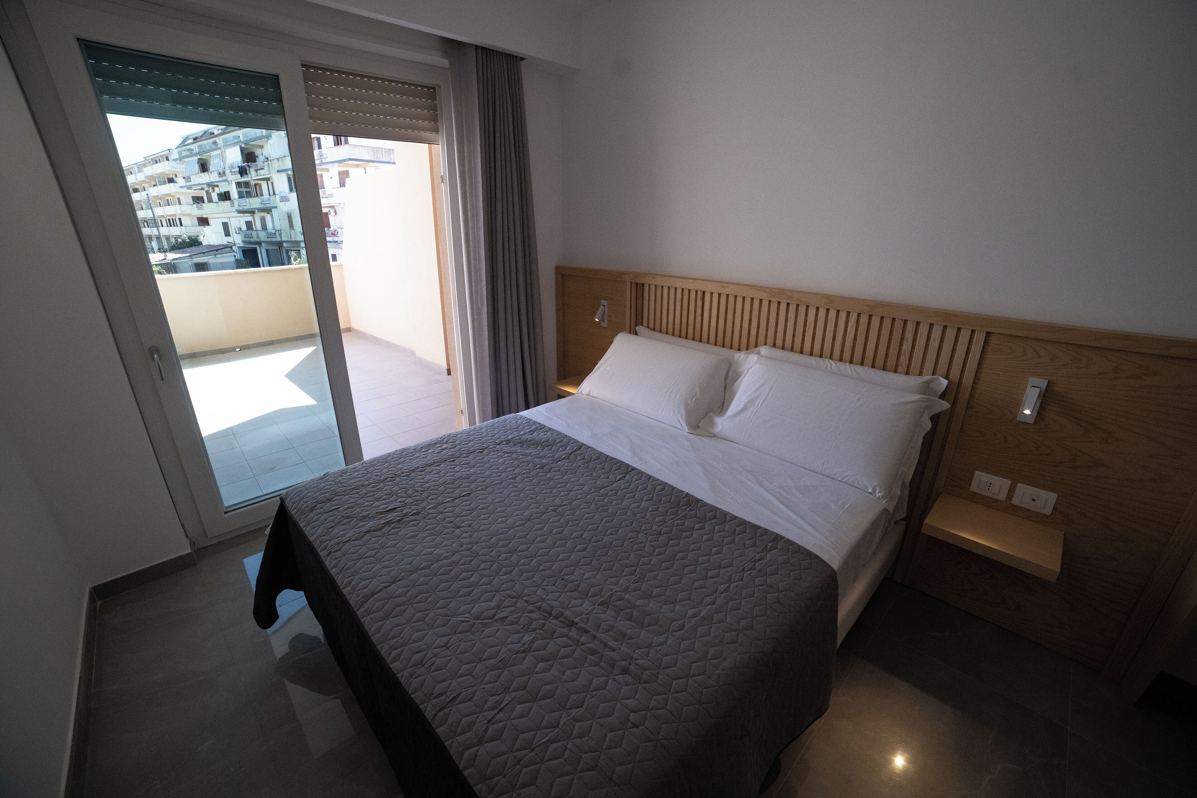 A hotel room with a bed, a wooden headboard, a small bedside table, and a view of the outside balcony.