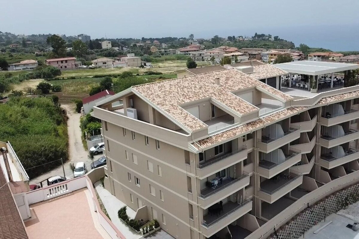 A multi-story residential building with balconies, a flat roof, and a view of a coastal town.