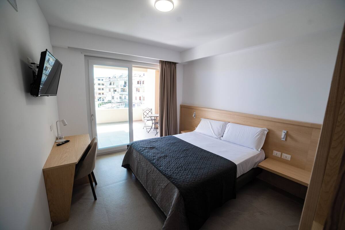 A hotel room with a bed, a desk, a chair, a TV, and a balcony with a view of buildings.
