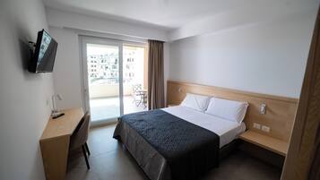 A hotel room with a bed, a desk, a chair, a TV, and a balcony with a view of buildings.