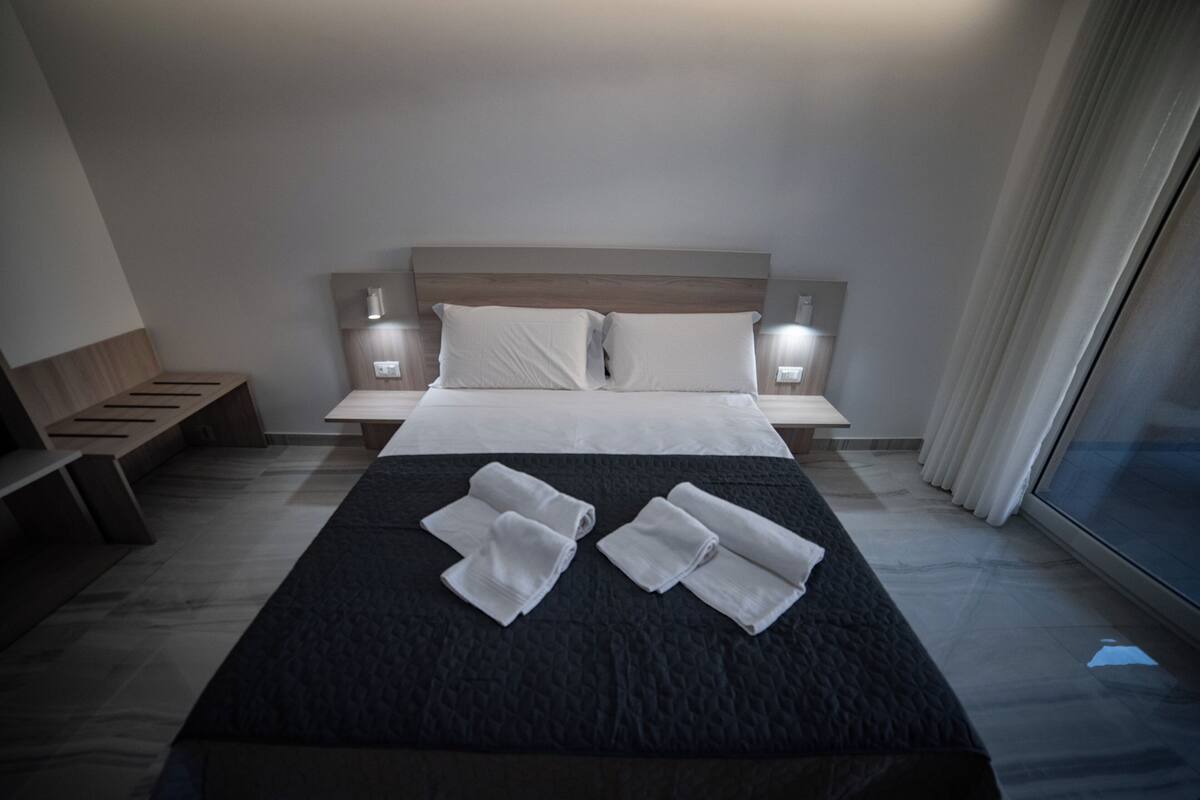 A hotel room with a large bed, two bedside tables with lamps, a wooden bench, and a view of the outside through a window.