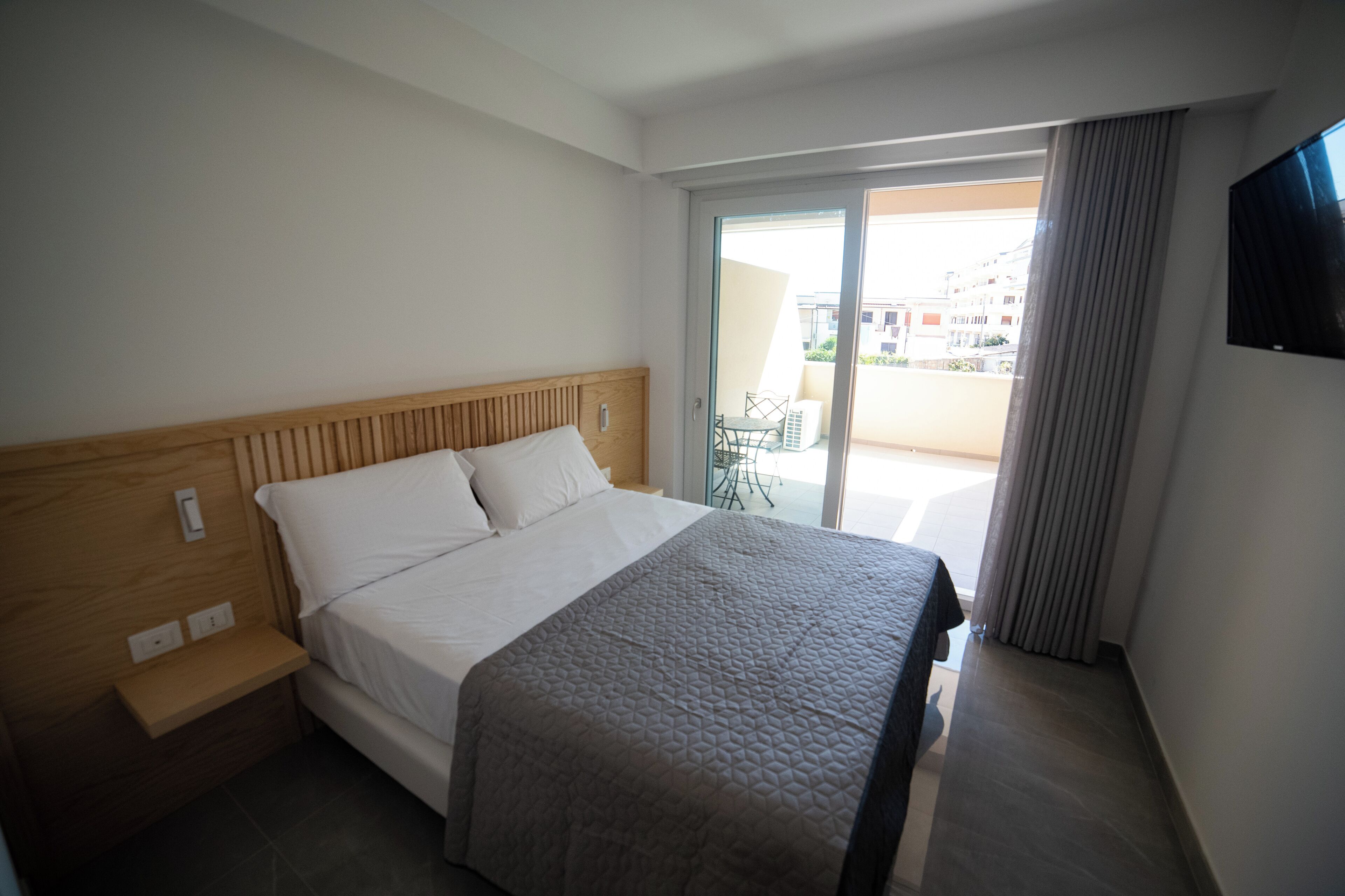 A hotel room with a bed, a wooden headboard, a bedside table, a TV mounted on the wall, and a sliding glass door leading to an outdoor area.