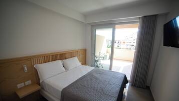 A hotel room with a bed, a wooden headboard, a bedside table, a TV mounted on the wall, and a sliding glass door leading to an outdoor area.
