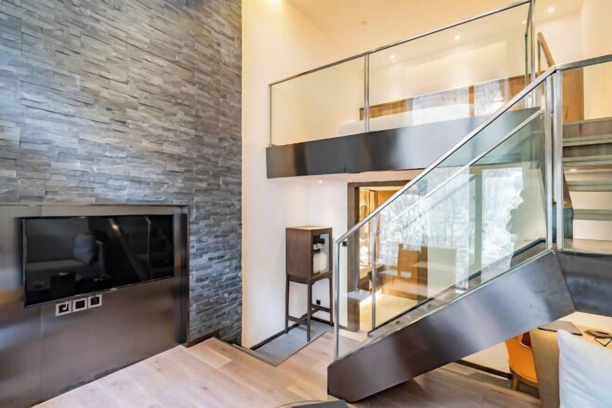 A modern interior with a stone wall, a flat-screen TV, a staircase with glass railings, and a dining area.