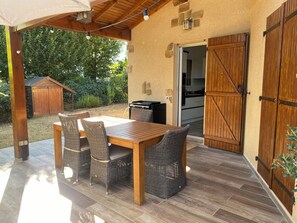 En uteplats med ett träbord och flätade stolar, en pergola med ljusslingor, en trädörr och ett litet förråd i bakgrunden.