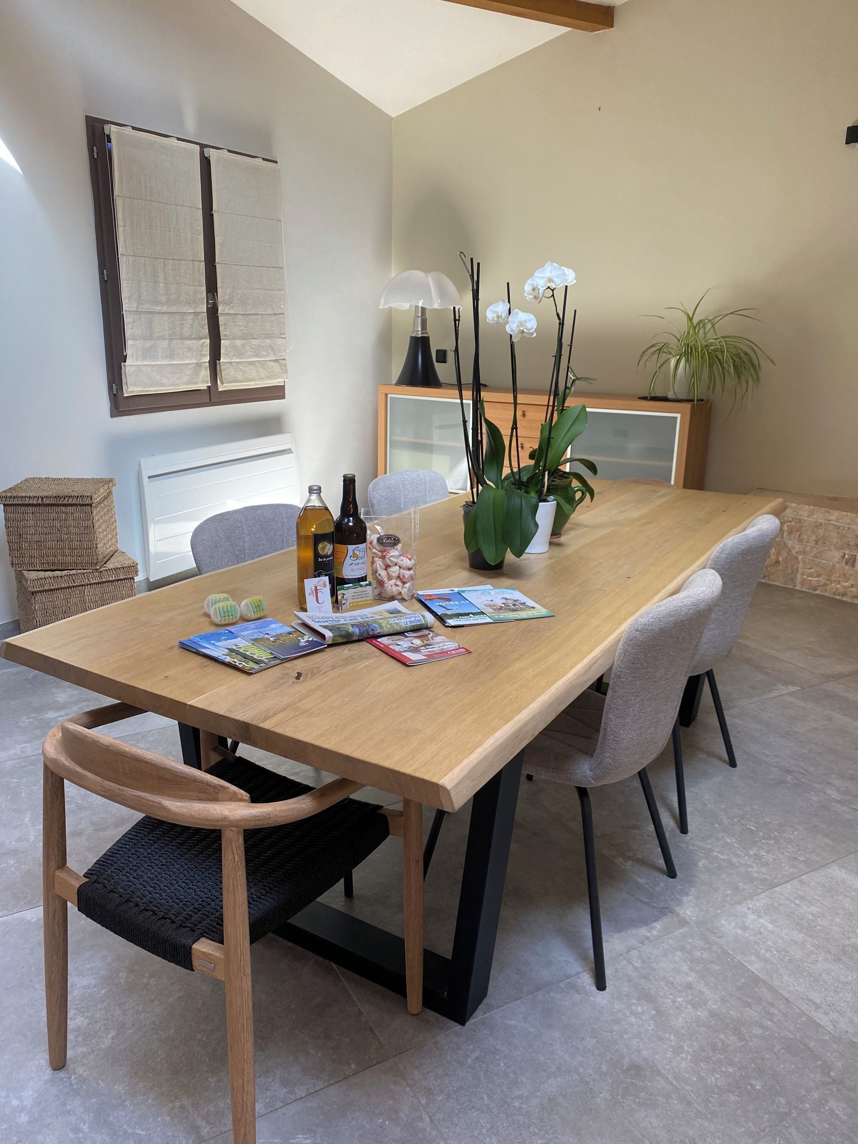 A dining area with a wooden table, grey upholstered chairs, a wicker basket, a potted plant, and a framed picture on the wall.