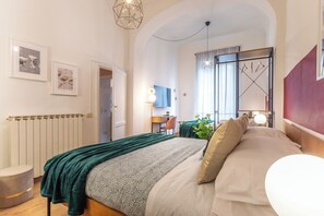 Un dormitorio moderno con una cama grande, un escritorio y una silla. Se observa un radiador, una planta y una ventana con cortinas.