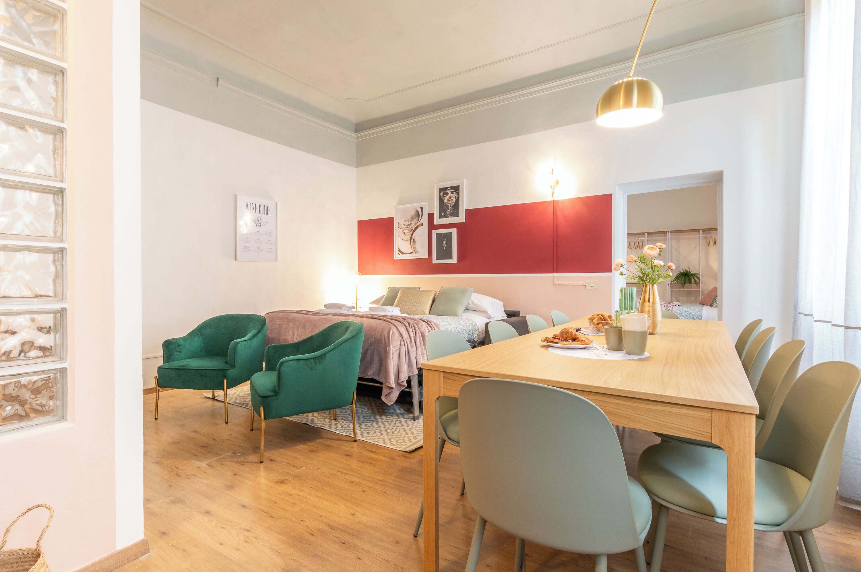 A modern living room with a wooden dining table, green chairs, a sofa, and a red accent wall.