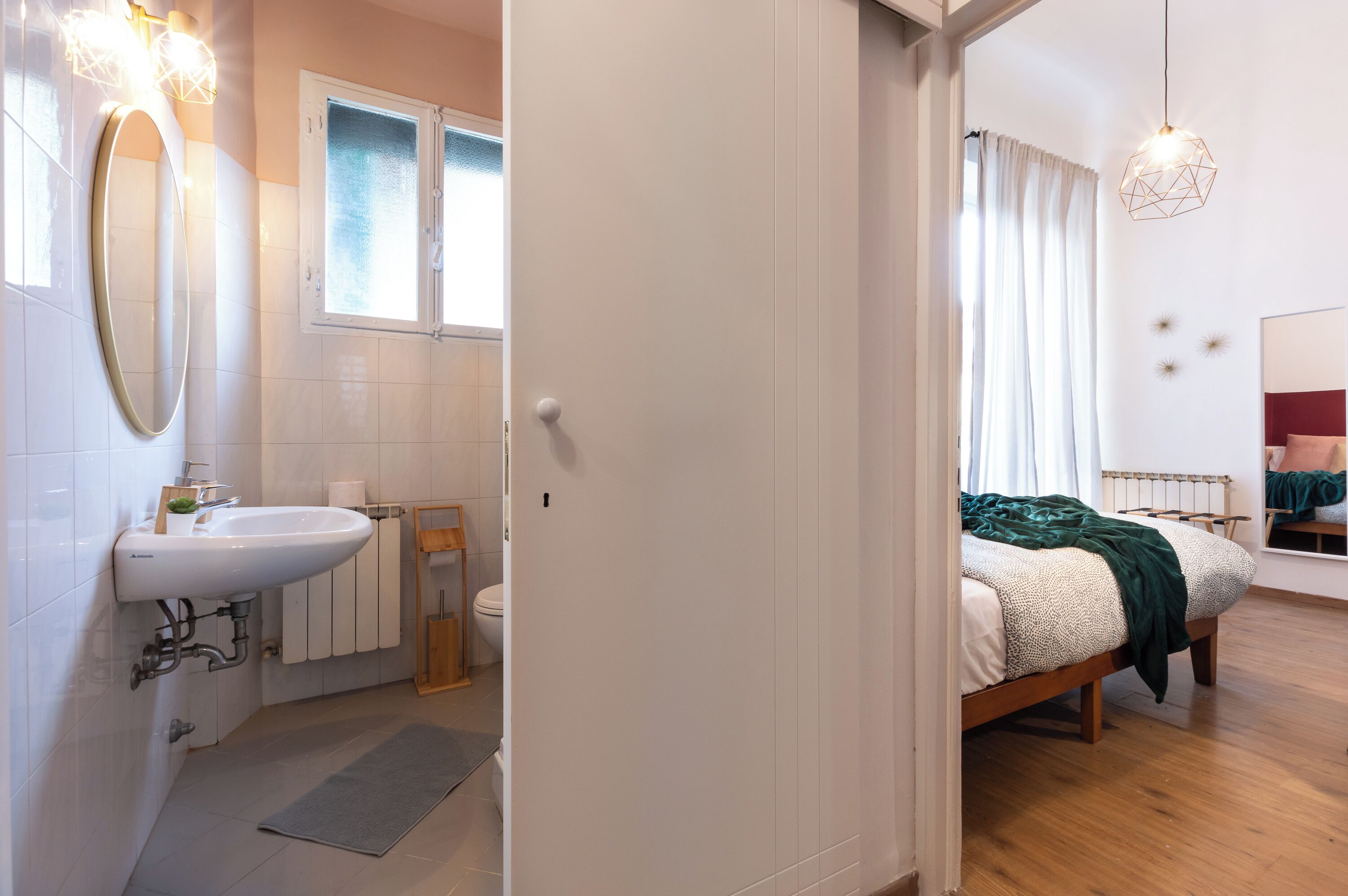 A modern bathroom with a sink, mirror, and window. The room has a wooden floor and a bed with a green blanket.