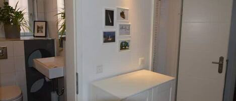 Una habitación pequeña y compacta con una puerta y un lavamanos blancos, y una pequeña estantería con fotografías enmarcadas.