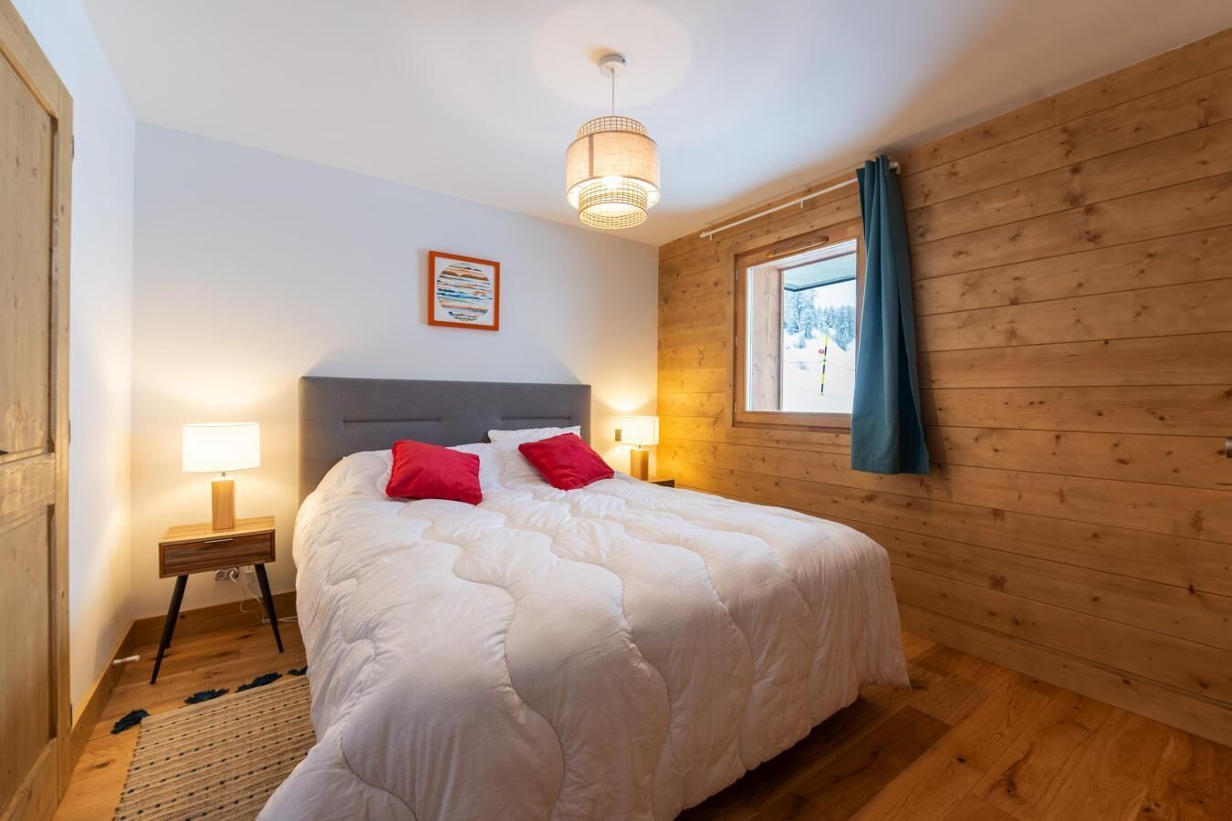 A bedroom with a large bed, wooden walls, a window with curtains, a bedside lamp, and a framed picture on the wall.