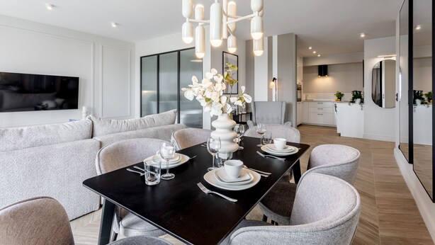 Un espace repas moderne avec une table noire dressée pour quatre personnes, entourée de chaises grises à revêtement. Un vase blanc rempli de fleurs est posé sur la table, et une grande fenêtre se trouve à l’arrière-plan.