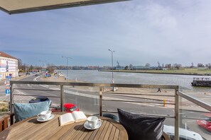 Een balkon met uitzicht op een rivier en een weg, een tafel met een boek en een kopje, en een rode auto die in de buurt geparkeerd staat.