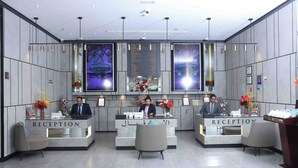 Un área de recepción con dos mostradores, cada uno atendido por un recepcionista. Hay arreglos florales sobre los mostradores y un panel informativo encima.