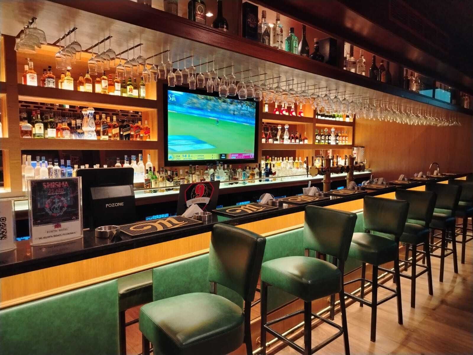 A bar area with a television screen, shelves of liquor bottles, and green bar stools.