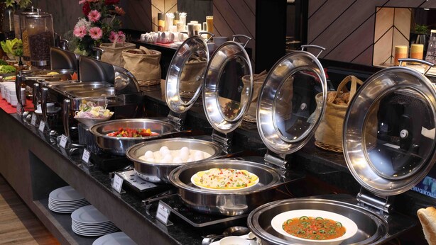 Montaje tipo bufé con diversos alimentos en recipientes cubiertos, platos apilados abajo y una estación de café.