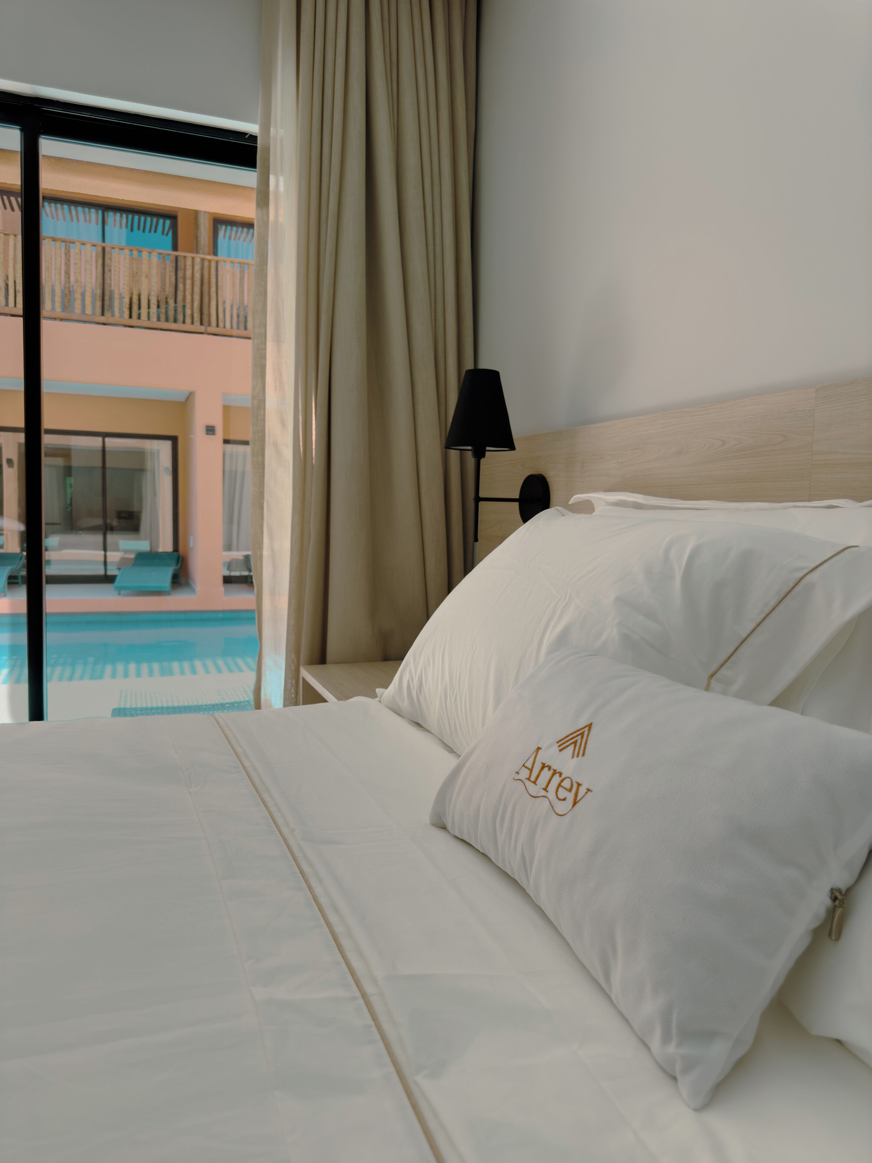 A hotel room with a bed, a nightstand, a lamp, and a view of a pool and balcony.