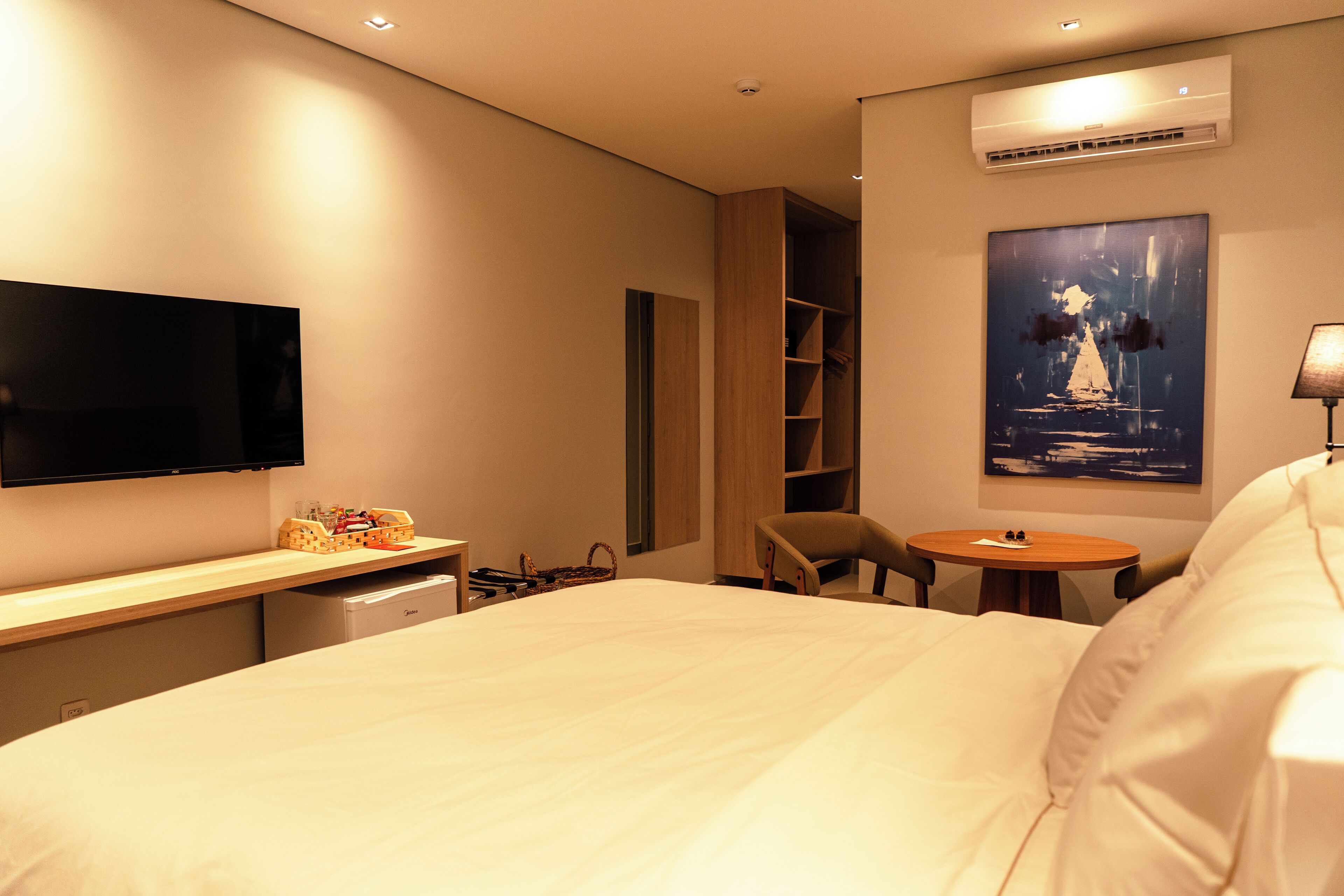 A hotel room with a large bed, a flat-screen TV mounted on the wall, a small desk with a lamp, a chair, and a painting on the wall.