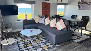 A modern living room with a grey sofa, a round coffee table, and a dining area with a table and chairs. A TV is mounted on the wall, and there is a view of the sea through the window. - The Lifeboat House, Skerries Harbour (Skerries)