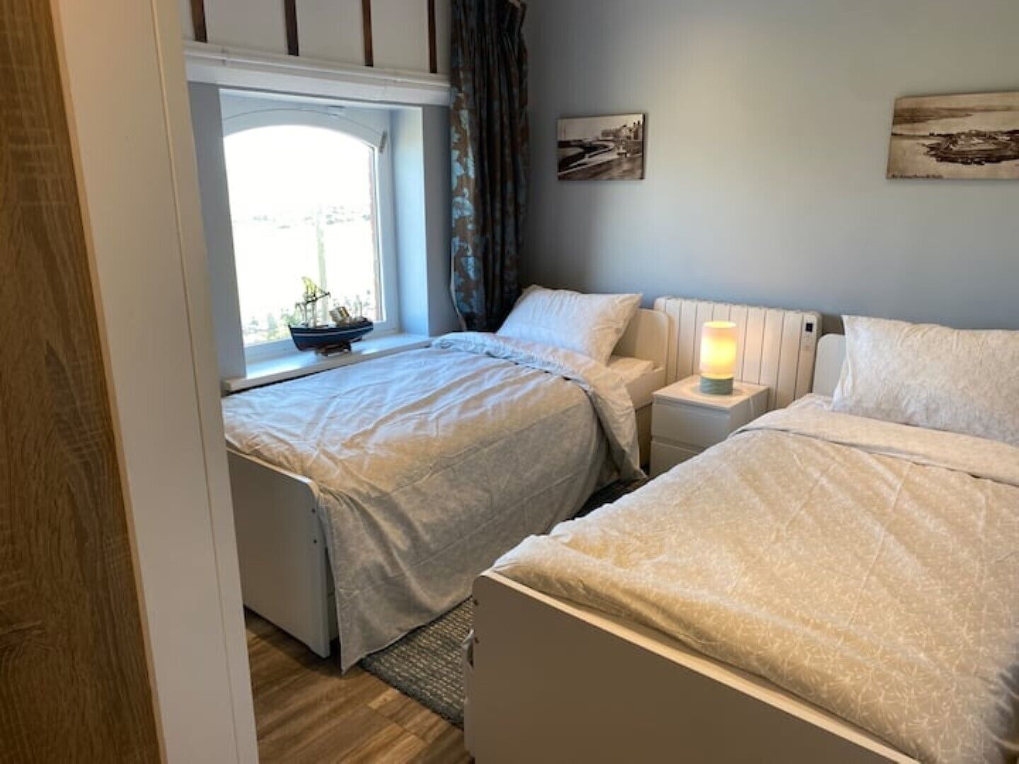 A bedroom with two beds, a window with a view, a bedside table with a lamp, and a small ship model on the windowsill.