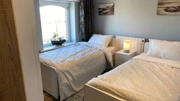A bedroom with two beds, a window with a view, a bedside table with a lamp, and a small ship model on the windowsill.