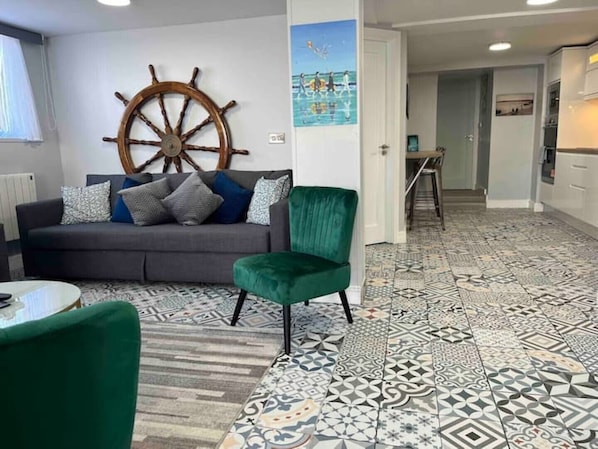 A living room with a grey sofa, green armchair, and a large wooden ship wheel decoration. The floor has patterned tiles.