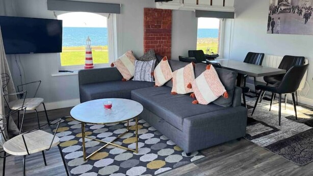 Ein modernes Wohnzimmer mit grauem Sofa, einem runden Couchtisch und einem Essbereich mit Tisch und Stühlen. Ein Fernseher ist an der Wand montiert und durch das Fenster ist Meerblick.