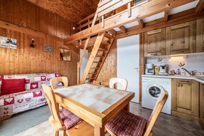 Una cabaña acogedora de madera con mesa, sillas, un sofá con cojines de estampado de corazón, una zona de cocina con lavadora y una escalera de madera.
