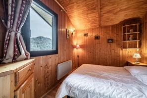 A wooden cabin room with a bed, a window with curtains, a bedside table, and a shelf with items. - Chalet, wifi, Ground floor, view mountain, parking, tv, 53m², Les Gets (Les Gets)