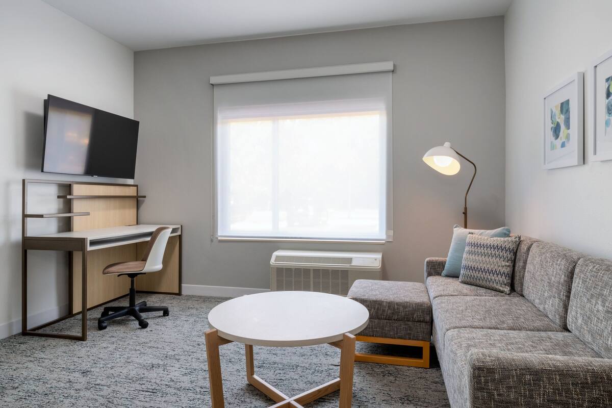 Ein modernes Wohnzimmer mit Flachbildfernseher, Schreibtisch, rundem weißen Tisch, grauem Sofa und Stehlampe.
