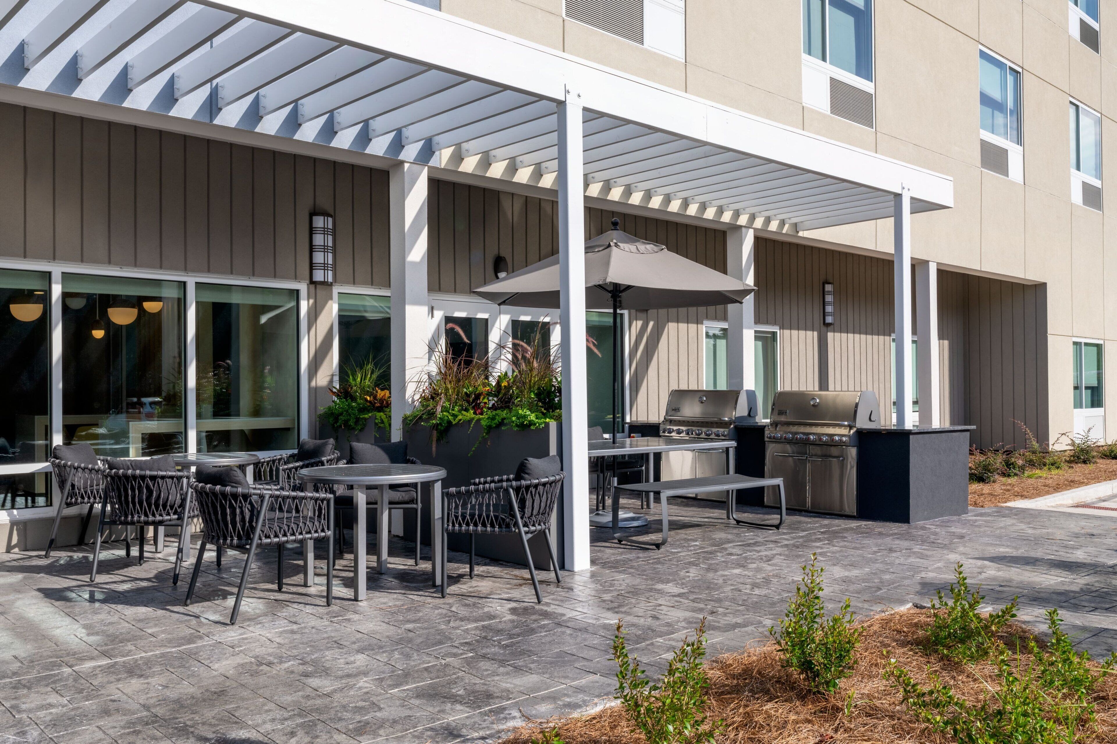 Een terras buiten met tafels, stoelen, een barbecue en een overdekt gedeelte met planten.