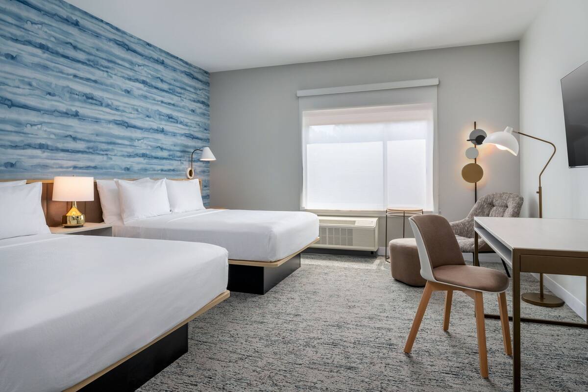 Ein Hotelzimmer mit zwei Betten, einem Schreibtisch, einem Stuhl, einem Fernseher und einem Fenster mit Jalousien.