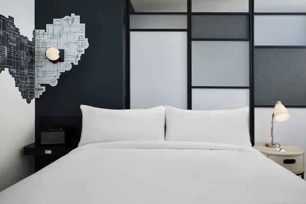 A modern hotel room with a neatly made bed, a nightstand with a lamp, and a decorative wall piece. - Aloft by Marriott Waukee (Waukee)