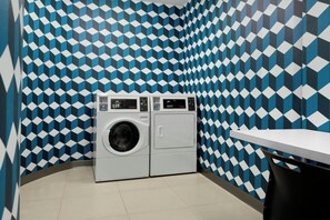 A laundry room with a patterned wall, a washing machine, and a table. - Aloft by Marriott Waukee (Waukee)