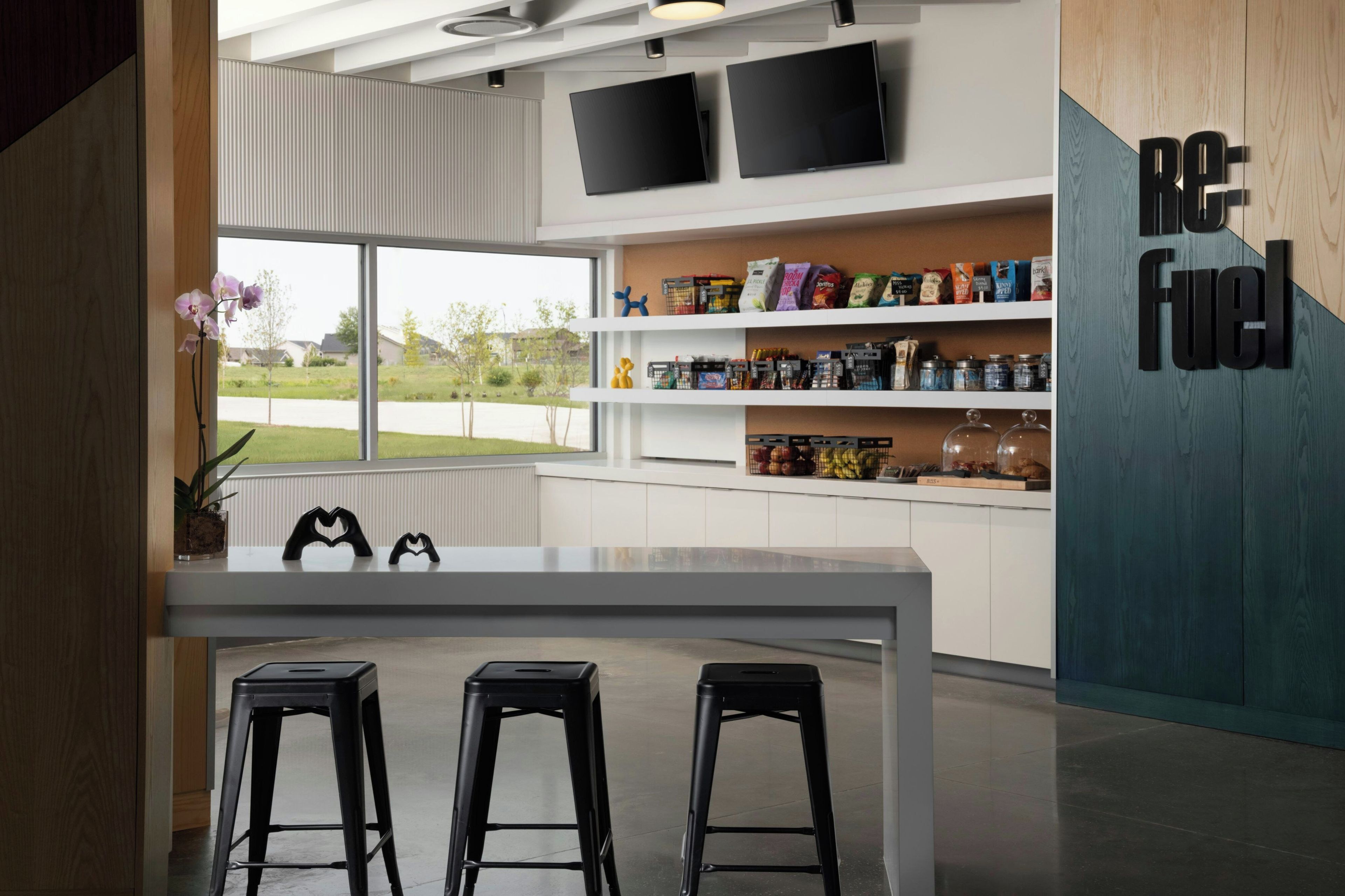 Una cocina moderna con encimera blanca, taburetes negros y vistas a una zona residencial a través de amplios ventanales.