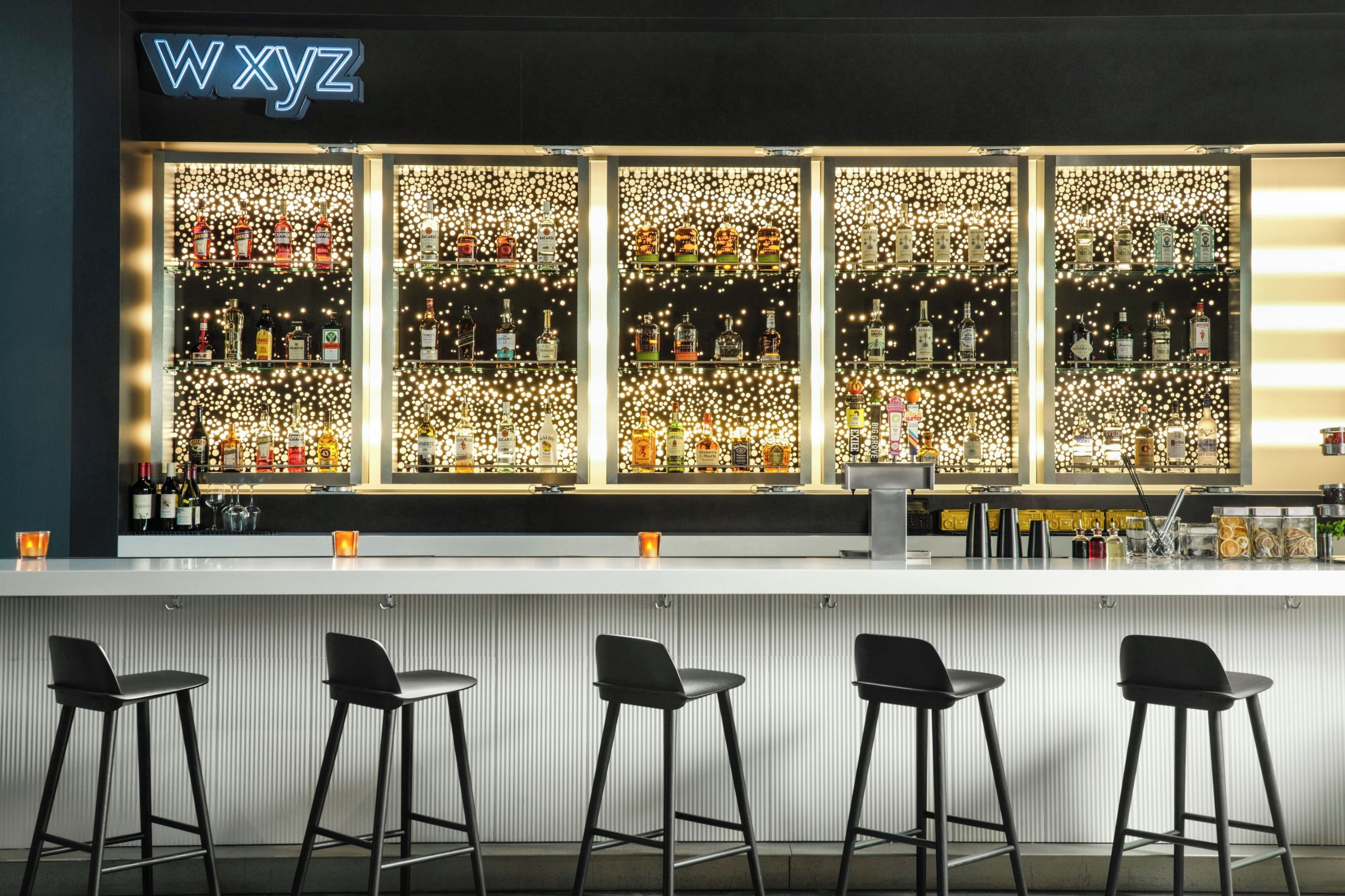 A bar area with a neon sign, multiple shelves of liquor bottles, and four bar stools.