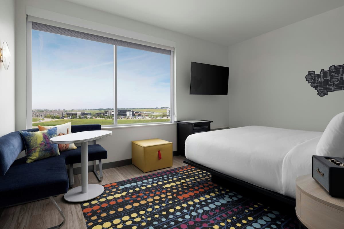 A modern hotel room with a large window, a bed, a small table, and a sofa.