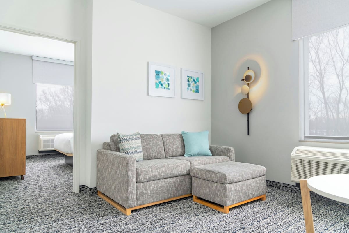 A modern hotel room with a grey sectional sofa, a round table, and a wall-mounted light fixture.