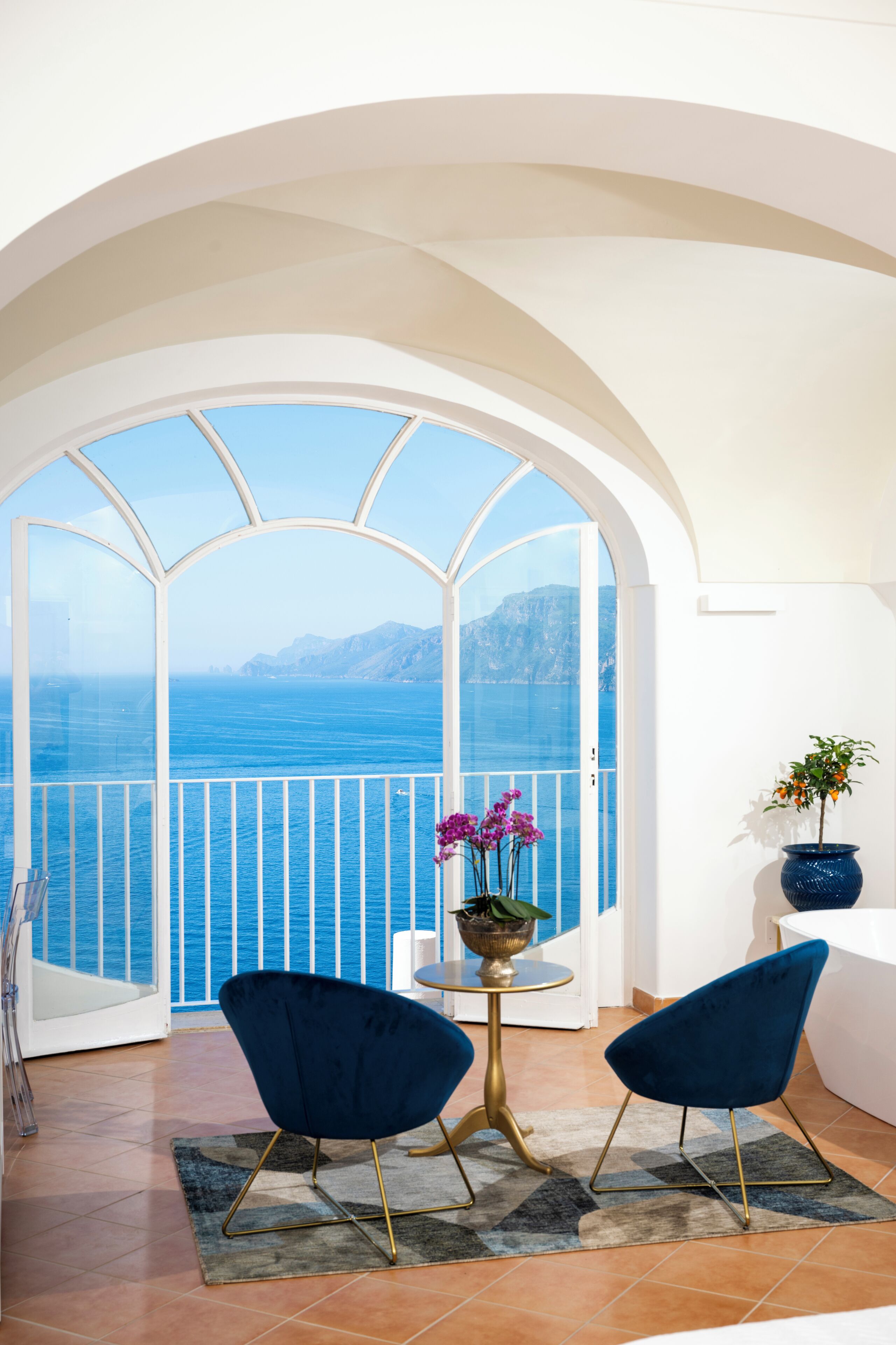Un balcone con vista sul mare, due sedie blu, un tavolino con una pianta e un vaso con fiori.