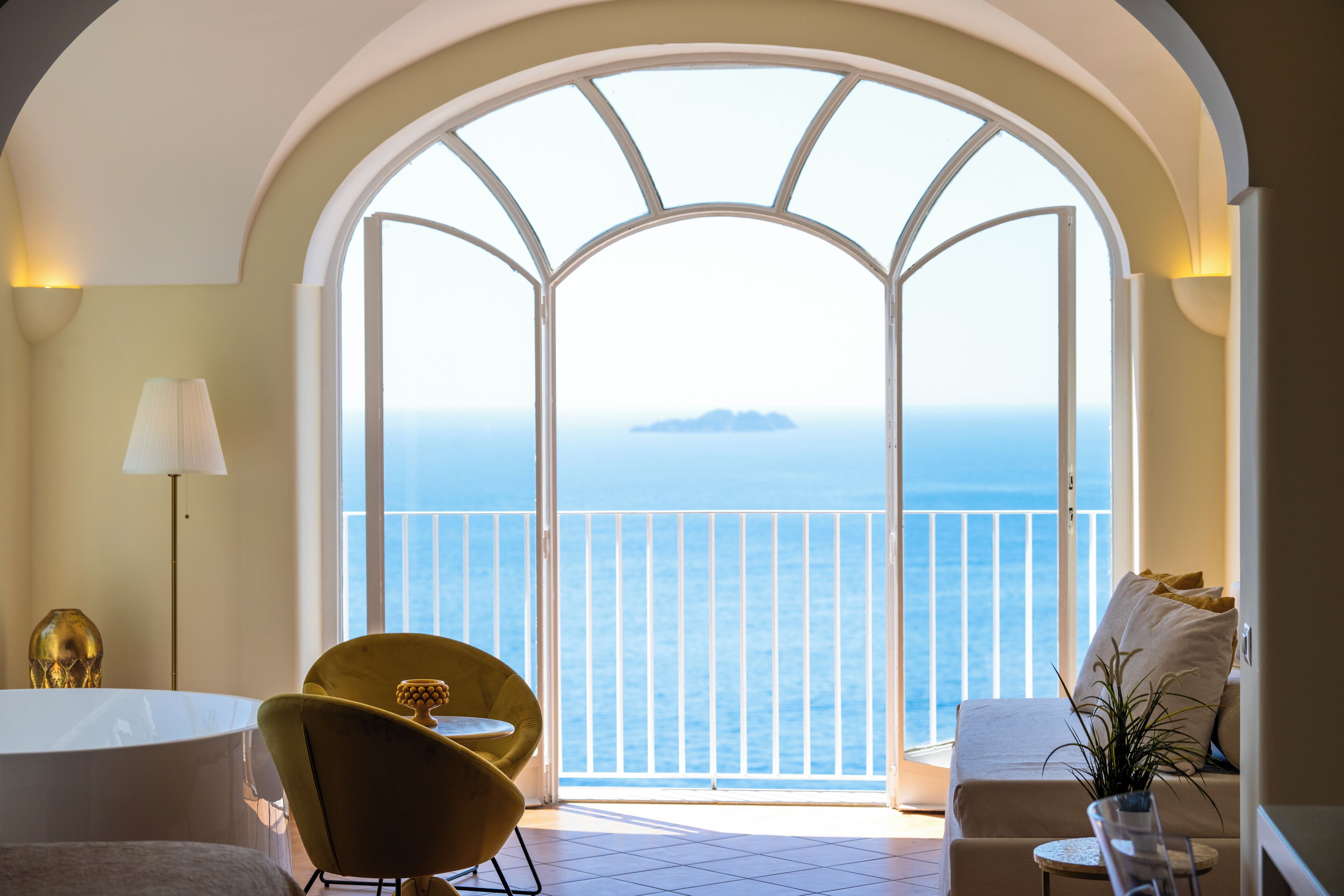 A room with a large arched window overlooking the sea, a round table, a yellow chair, and a sofa with a plant.