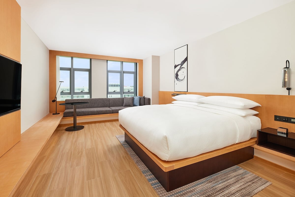 A modern hotel room with a large bed, a sofa, a TV, and a window with a city view.