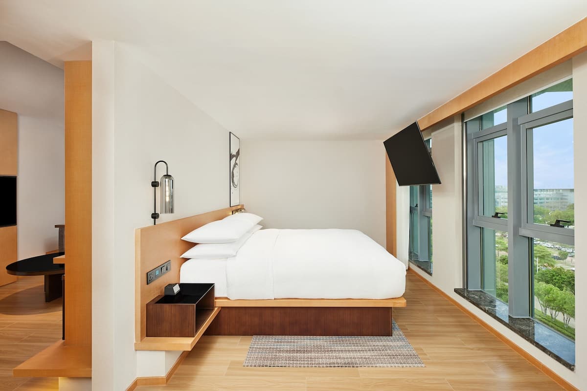 A modern bedroom with a bed, bedside tables, a flat-screen TV, and large windows offering a city view.