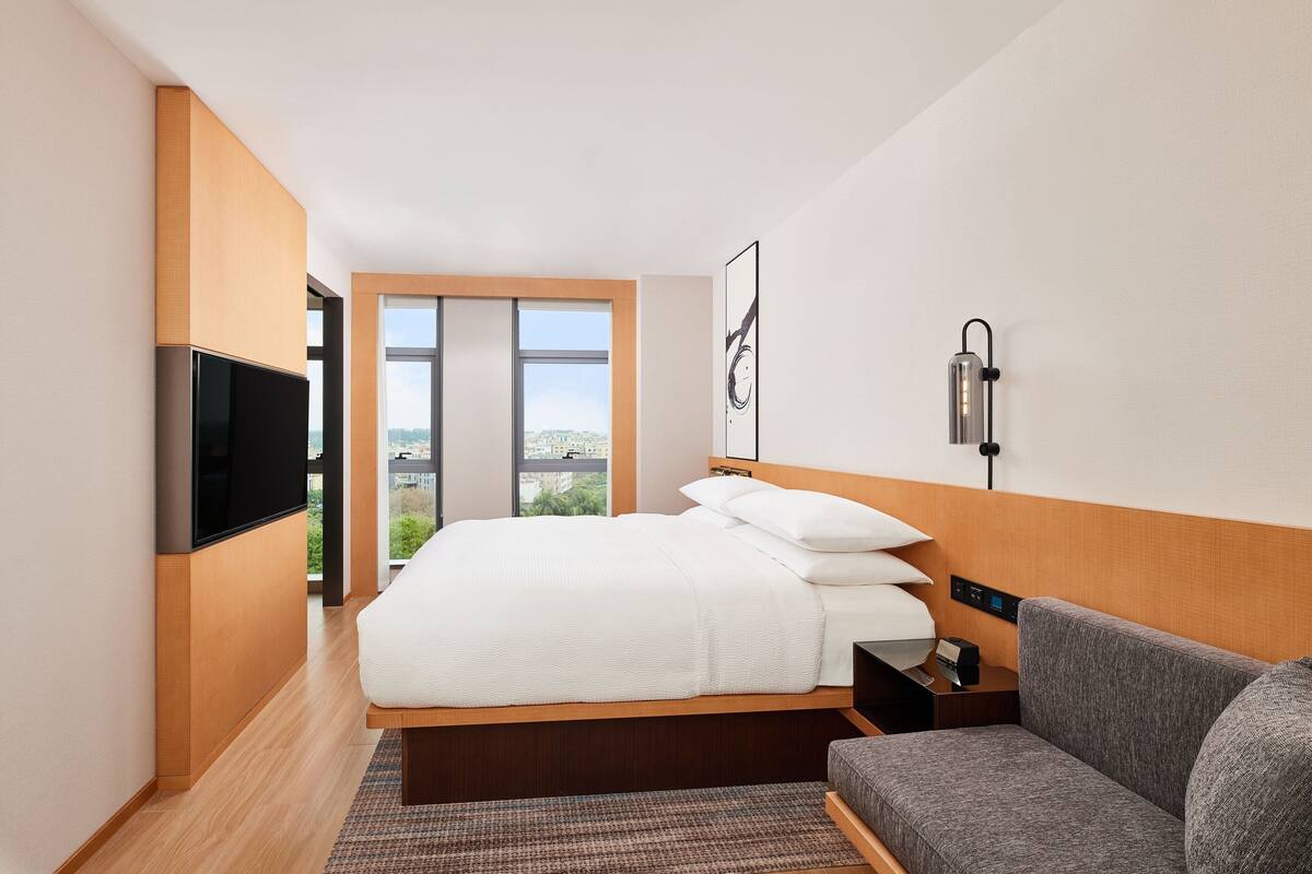 A modern hotel room with a large bed, a flat-screen TV, a bedside table, a gray sofa, and a large window offering a city view.
