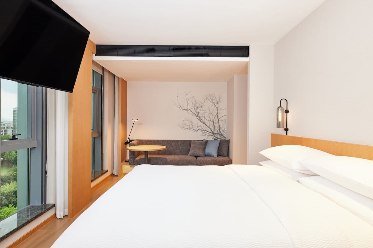 A hotel room with a large bed, a sofa, a small round table, and a view of the city through large windows.