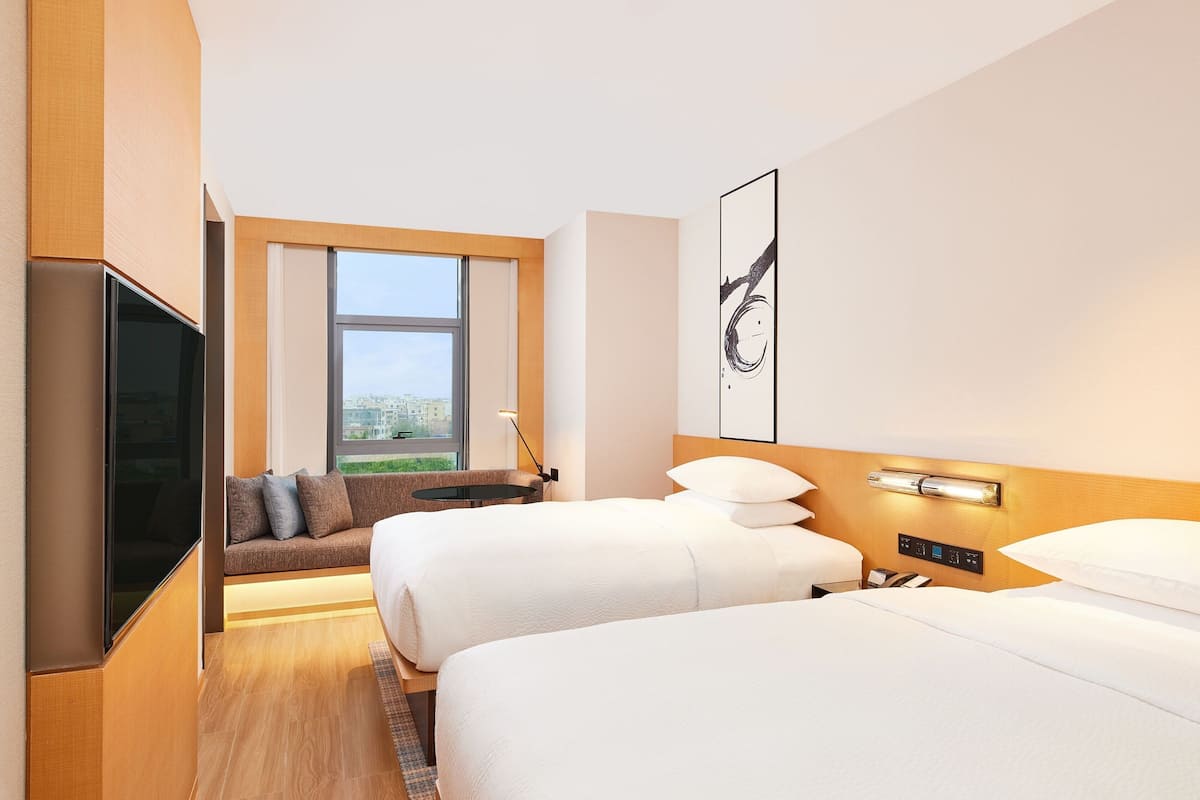 A hotel room with two beds, a sofa, a TV, a window with a view, and a wall-mounted artwork.