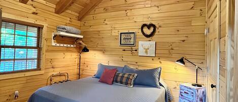 A wooden cabin bedroom with a bed, a ceiling fan, a window, and a wall-mounted heater.