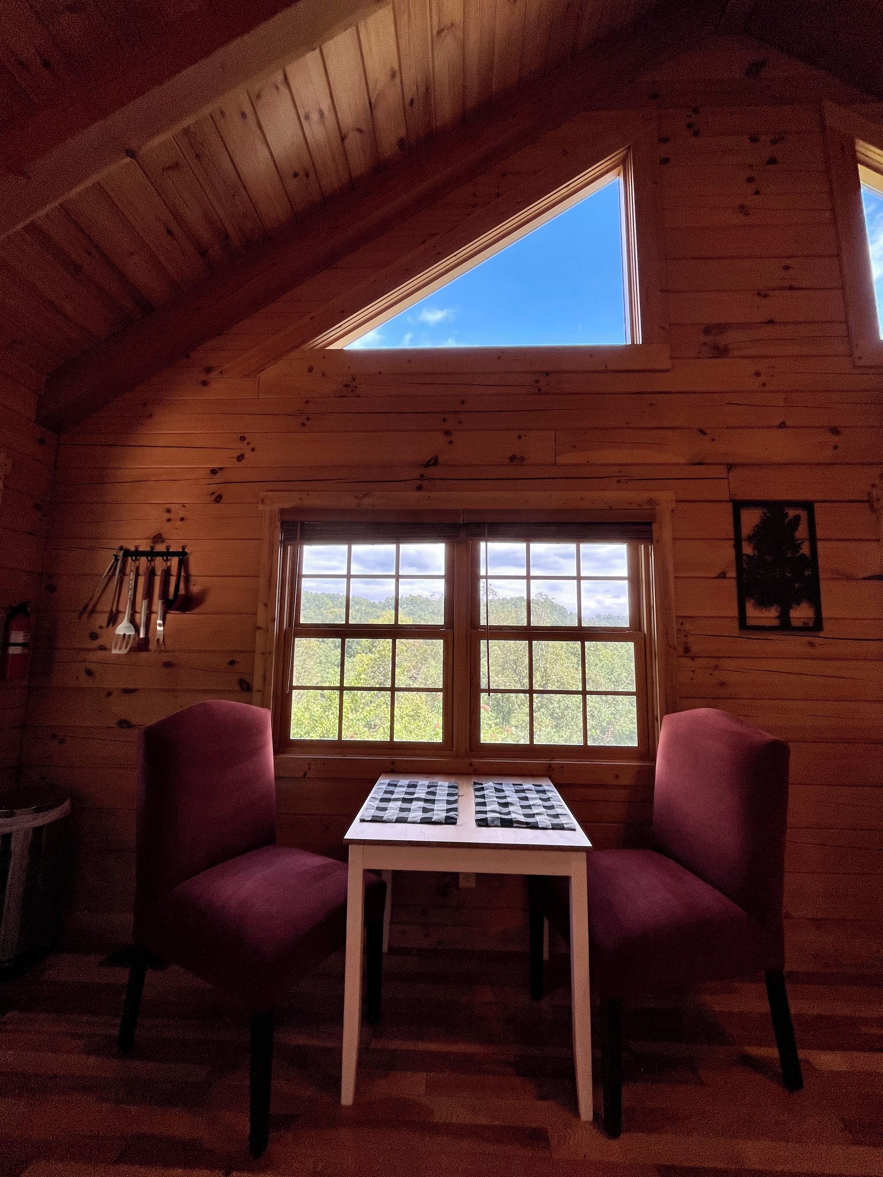 A cozy cabin room with a sloped ceiling, two purple chairs, a small table, and a large window offering a view of the outdoors.