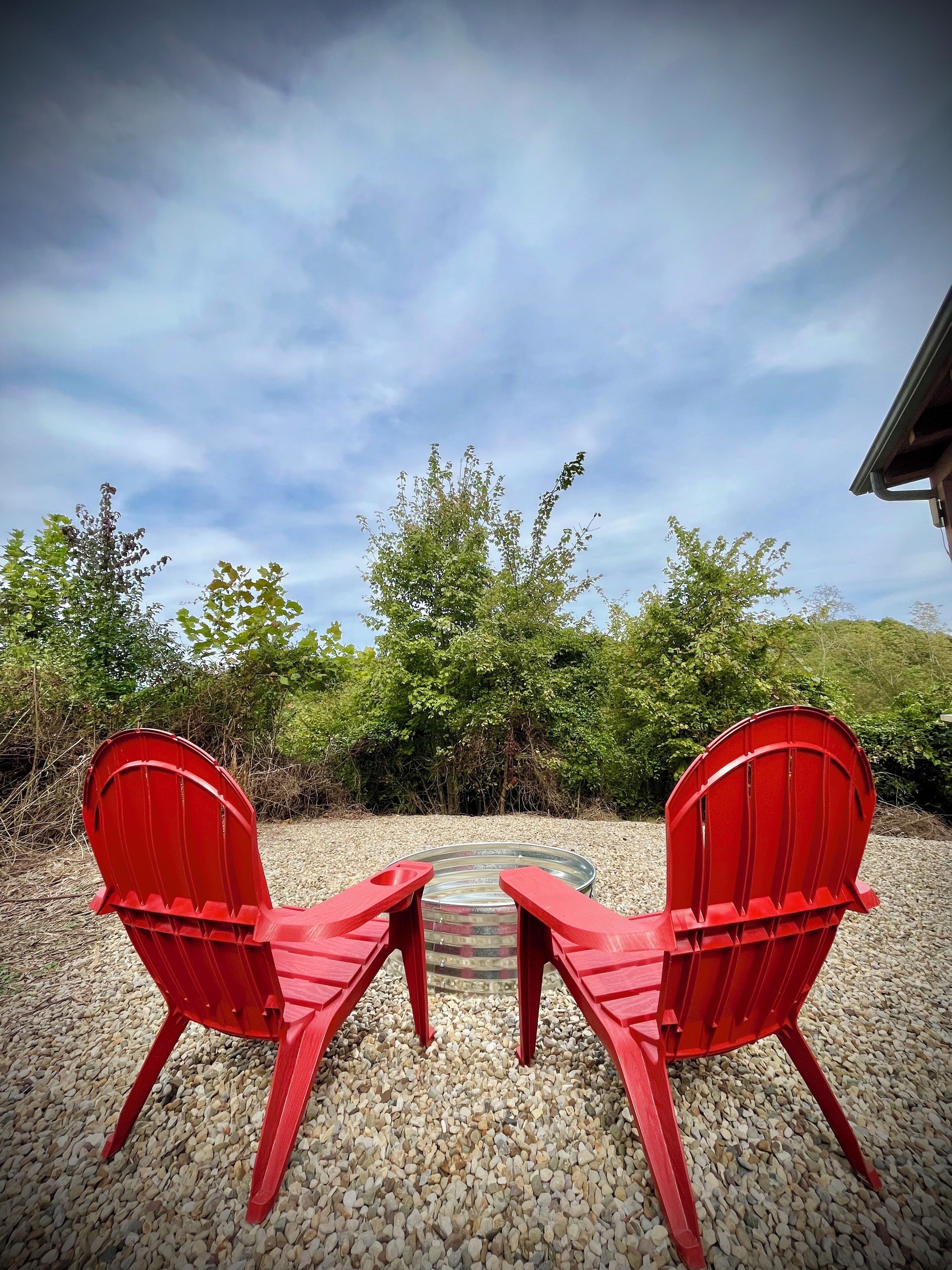 Two red Adirondack chairs facing each other with a gravel ground and greenery in the background.